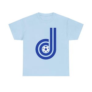 Puede incluir: Camiseta azul claro con un diseño azul de una letra "D" estilizada con un balón de fútbol. El diseño utiliza círculos concéntricos y líneas verticales, dando un aspecto retro. Una camiseta informal para los aficionados al deporte.