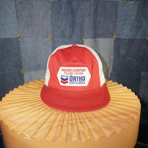 Puede incluir: Gorra de camionero roja y blanca con visera roja y parte trasera de malla blanca. La parte delantera de la gorra tiene un parche con el texto "MOORE AVIATION TULARE-COLUSA Chevroc ORTHO FERTILIZERS". La gorra está sobre una superficie texturizada marrón claro.