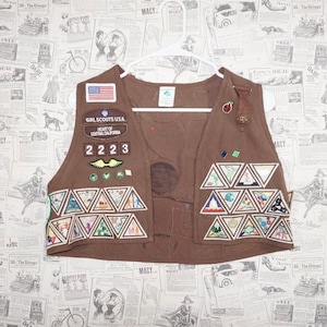 May include: A brown Girl Scout vest with various patches and badges. The vest features an American flag patch, a patch that says "Girl Scouts USA Heart of Central California", and the number "2223". The vest is decorated with multiple triangular patches.