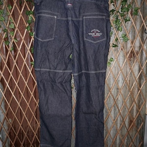 May include: Dark blue denim jeans with white stitching and a patch on the back pocket. The patch has text that reads "SINGAPORE WILLIE BSCO ARA FASHION". The jeans have a classic five-pocket design.
