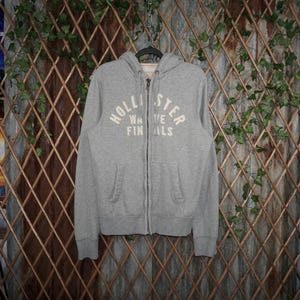 May include: A gray zip-up hoodie with the text "HOLLISTER WAVE FINALS" in white across the chest. The hoodie has a drawstring hood and two front pockets. It is hanging on a black hanger.