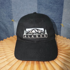 May include: Black baseball cap with a white embroidered design featuring mountains, trees, and the word "ALASKA" in a rectangular frame. The cap has a curved brim and a classic six-panel design.