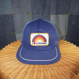 Op de afbeelding: Blauwe truckerpet met een mesh achterkant en een gebogen klep. De voorkant heeft een patch met een regenboogontwerp en de tekst "I COZUMEL". De rand is omrand met witte stiksels.