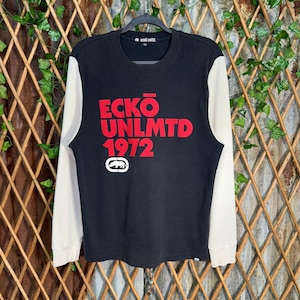 May include: Black long-sleeve sweatshirt with cream-colored sleeves. The front features the red text "ECKO UNLMTD 1972" and a white logo. The sweatshirt is hanging on a black hanger against a backdrop of wooden slats and greenery.