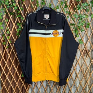 May include: Black, yellow, and white zip-up jacket with the Los Angeles Lakers logo. The jacket has a black collar, sleeves, and back. The front features a yellow lower section, a white stripe, and the Lakers logo.
