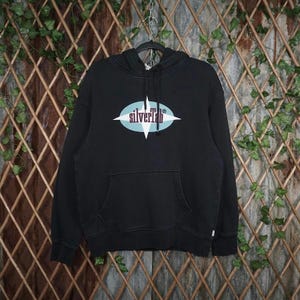 May include: Black hooded sweatshirt with a front pocket and the text "silverTab" in a white and light blue design. The sweatshirt is hanging on a black hanger against a backdrop of wooden lattice and greenery.