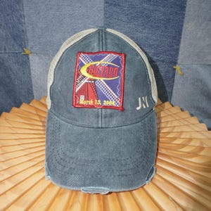 May include: A distressed blue baseball cap with a white mesh back. The front features a patch with the text "Sarvang Family Park" and the date "March 13, 2004". The cap has a curved brim and a small embroidered "JN" on the side.