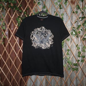 May include: Black crew neck t-shirt with a grey and beige graphic design. The graphic features an eagle, the year 1989, and floral elements. The shirt has short sleeves and a grey neckline.
