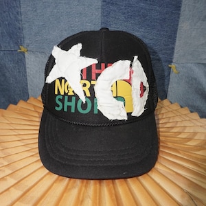 May include: Black trucker hat with a white star and a white cutout design over the text "THE NORTH SHORE" in red, yellow, green, and black. The hat has a curved brim and a mesh back.