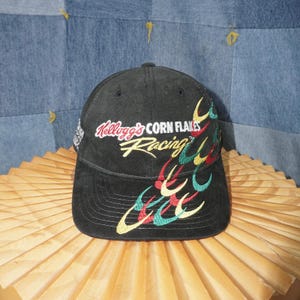 May include: Black baseball cap with the text "Kellogg's Corn Flakes Racing" embroidered in yellow, red, and green. The cap has a curved brim and a structured crown. The cap is sitting on a light brown surface.