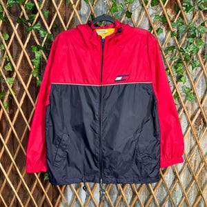May include: A red and navy blue zip-up jacket with a hood. The jacket features a white stripe across the chest and a small logo on the chest. The jacket is hanging on a black hanger.
