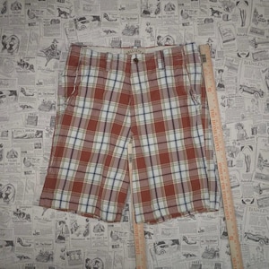 May include: Red, white, and blue plaid shorts with a button closure. The shorts have pockets and a classic plaid pattern. A wooden ruler is placed next to the shorts. The shorts are made by Hollister.