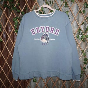 May include: A blue-grey sweatshirt with a graphic of Eeyore from Winnie the Pooh. The word "Eeyore" is in purple block letters above an image of Eeyore. The text "Another Gloomy Day" is below the image.