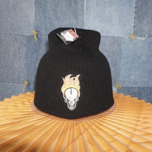 May include: Black knit beanie with a skull and flame patch. The patch features a white skull with yellow and orange flames. A tag is attached to the beanie. The beanie is displayed on a wooden surface.