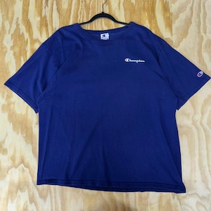 Puede incluir: Una camiseta Champion azul marino con mangas cortas. El logotipo de Champion está bordado en blanco en el pecho y en el puño de la manga izquierda. La camiseta está colgada de una percha negra sobre un fondo claro de vetas de madera.