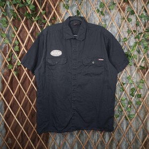 May include: A black short-sleeve button-down shirt featuring a classic collar and two chest pockets. The right chest pocket displays an oval patch with the text "DICKIES" in white, while the left pocket features a small rectangular brand label.