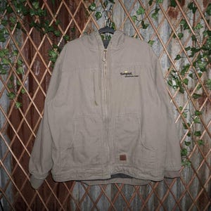 Può includere: Giacca con cappuccio beige con chiusura a zip completa, due tasche frontali e un piccolo logo sul petto. La giacca è appesa a una gruccia di metallo su uno sfondo di traliccio di legno con viti verdi.