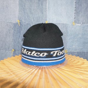 Può includere: Berretto nero a maglia con la scritta "Matco Tools" in bianco, bordata da strisce blu e bianche. Il bordo è blu. Il cappello è esposto su una superficie marrone chiaro.