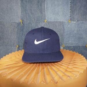 Op de afbeelding: Marineblauwe baseballcap met een witte Nike swoosh-logo. De cap heeft een gestructureerde pasvorm en een gebogen klep. De hoed wordt gepresenteerd op een lichtbruin, getextureerd oppervlak.