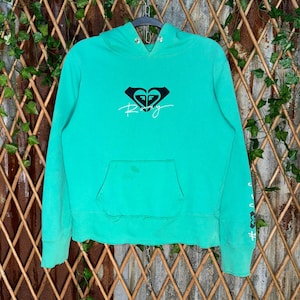 May include: A turquoise hoodie with a front pocket and a hood. The hoodie features a black and white logo with the word "Roxy" in cursive. The sleeve has a white graphic. The garment is hanging on a metal hanger.