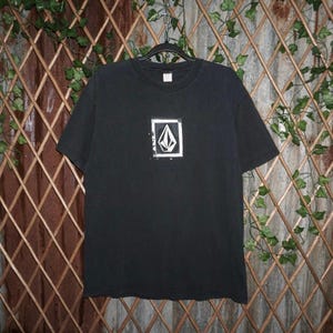 May include: Black short-sleeved t-shirt with a white square logo on the front. The logo features a stylized, geometric design. The shirt is hanging on a black hanger against a brown and green background.
