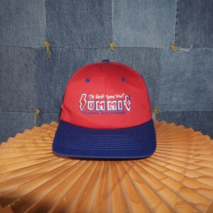 May include: A red and blue baseball cap with the word "SUMMIT" in white and blue lettering. The text "The World's Speed Shop" is above the word "SUMMIT". The cap has a curved brim and a button on top.