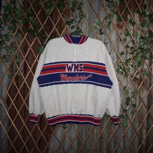 May include: A grey sweatshirt with a quarter-zip neckline. The front displays the text "WMS Warriors" in red and blue stripes. The cuffs and collar have matching red and blue striped trim. The sweatshirt is hanging on a black hanger.