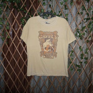 Könnte beinhalten: Beige T-Shirt mit Grafik einer Person, die Gitarre spielt, mit dem Text "Crosby Stills Nash & Young" und dem Namen der Band. Das T-Shirt hängt an einem schwarzen Kleiderbügel vor einem dekorativen Hintergrund.