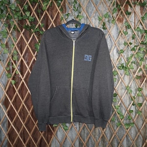 May include: A charcoal gray zip-up hoodie with a blue interior lining and a yellow zipper. The hoodie features a blue and white logo on the chest. The hoodie is hanging on a black hanger against a wooden trellis background.