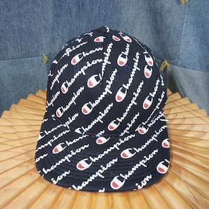 May include: Navy blue baseball cap with a repeating white "Champion" logo and red accents. The hat features a curved brim and a classic six-panel design, suitable for casual wear.