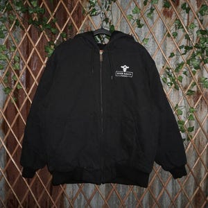 May include: Black hooded zip-up jacket with the "River Ranch Farms" logo on the upper left chest. The jacket has a full zipper, a hood, and ribbed cuffs and hem. The jacket is hanging on a black hanger.