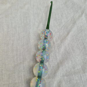 May include: A crochet hook with a green handle and a series of four clear, round beads. The beads are decorated with colorful confetti-like dots. The hook is designed for crafting and textile work.