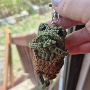 May include: A crocheted frog keychain featuring a dark olive green body with black bead eyes and brown yarn overalls. The small stuffed figure is attached to a silver-toned metal lobster clasp, showcasing detailed yarn stitching and texture.