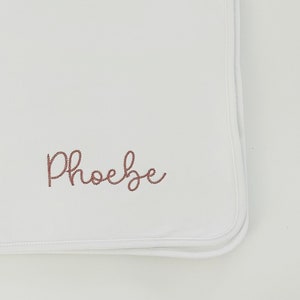 May include: White baby blanket with a pink embroidered name "Phoebe" on it.