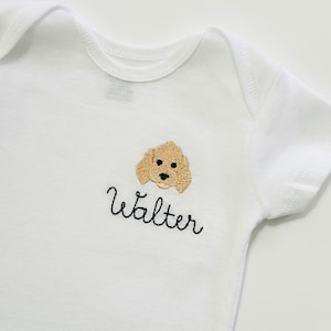 May include: White baby bodysuit featuring a tan embroidered dog face and the name "Walter" in black script. The garment has short sleeves and a round neckline. The design is on the chest.