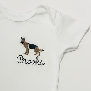 Puede incluir: Uniforme blanco para bebé con un pastor alemán bordado y el nombre "Brooks" debajo.