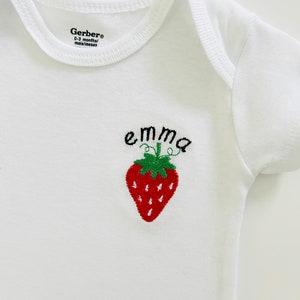 Puede incluir: Un body blanco para bebé con una fresa bordada en rojo y el nombre "emma" en bordado negro.