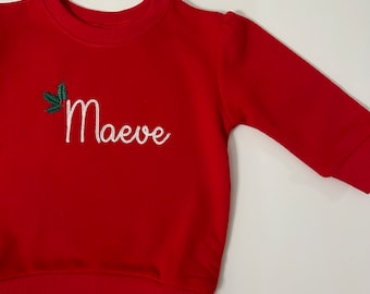 May include: Red long-sleeve sweatshirt with a white embroidered name "Maeve" and a green holly leaf design.