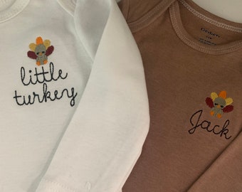 May include: Two long-sleeved baby onesies. One is white with a brown turkey and the text "little turkey". The other is brown with a brown turkey and the text "Jack".