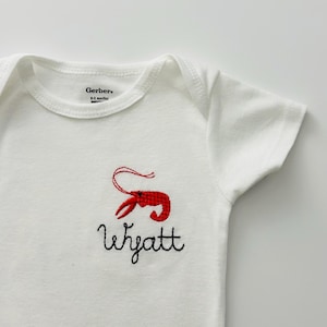 Peut inclure: Combinaison blanche pour bébé avec un homard rouge et le nom "Wyatt" brodé sur le devant. Marque Gerber, 0-3 mois.