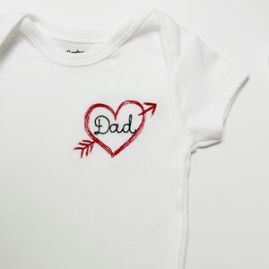 May include: White baby onesie with a red embroidered heart with an arrow through it. The word "Dad" is embroidered inside the heart in black thread.