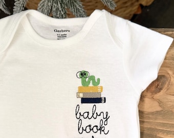 May include: White baby onesie with a green worm wearing glasses embroidered above a stack of books. The text "baby book worm" is embroidered below the books. The onesie is labeled "Gerber's".