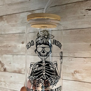 May include: A clear glass reusable water bottle with a bamboo lid and a black and white graphic of a skeleton with a messy bun, sunglasses, and the text "Dead Inside But Caffeinated".