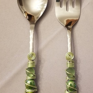 May include: A set of two stainless steel serving utensils with green glass beads wrapped around the handles. The utensils are a spoon and a fork.
