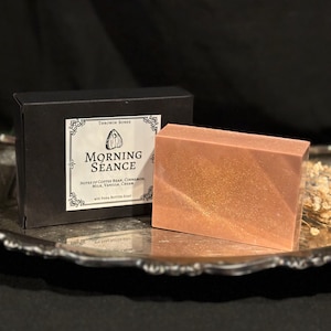 Puede incluir: Una barra de jabón de manteca de karité rosa con purpurina dorada. El jabón está empaquetado en una caja negra con una etiqueta blanca que dice "Throwin Bones Morning Séance Notes of Coffee Bean, Cinnamon, Milk, Vanilla, Cream 4oz Shea Butter Soap".