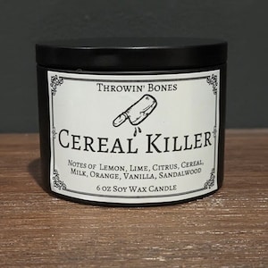 May include: Black candle jar with a white label that reads "THROWIN' BONES CEREAL KILLER" with a drawing of a butcher knife. The label also lists the scent notes: "NOTES OF LEMON, LIME, CITRUS, CEREAL, MILK, ORANGE, VANILLA, SANDALWOOD" and "6 OZ SOY WAX CANDLE".