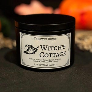 May include: A black 6 oz soy wax candle in a metal tin. The label reads "Throwin' Bones Witch's Cottage" with a witch hat illustration. Scent notes include buttery pecan, sweet pumpkin, cinnamon, vanilla, and maple syrup.