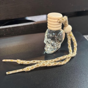 Puede incluir: Una botella transparente con forma de calavera de cristal con tapón de madera y cuerda decorativa. La cuerda es beige con detalles dorados. La botella es probablemente un ambientador o un objeto decorativo.