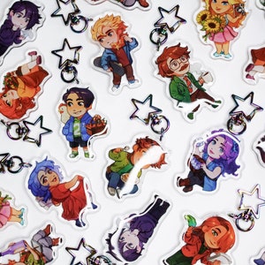 May include: A collection of colourful cartoon character keychains with star-shaped charms. The characters are drawn in a cute anime style and feature a variety of outfits and accessories.