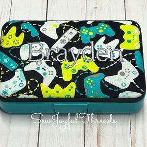 May include: A teal blue rectangular storage box with a black lid. The lid is covered in a black fabric with a repeating pattern of white, yellow, and blue video game controllers. The name "Brayden" is embroidered in white on the lid.